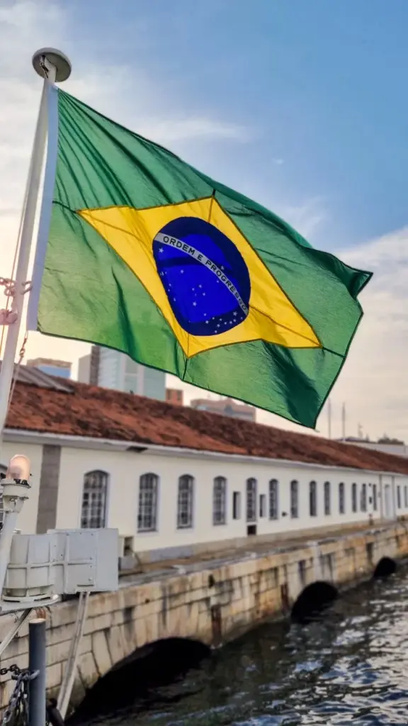 Brazilian flag waving near a historic building by the water in Rio de Janeiro, creating a vibrant urban scene.