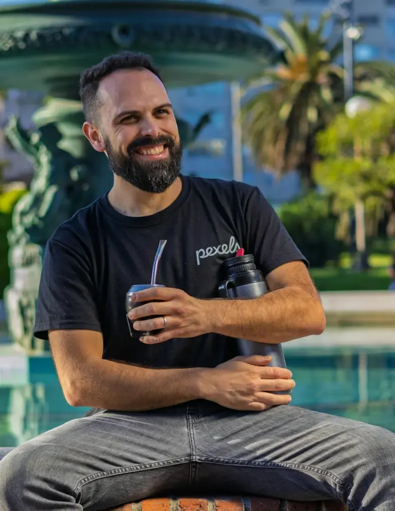 Bearded man sitting by a fountain, holding a traditional mate and thermos, smiling warmly.