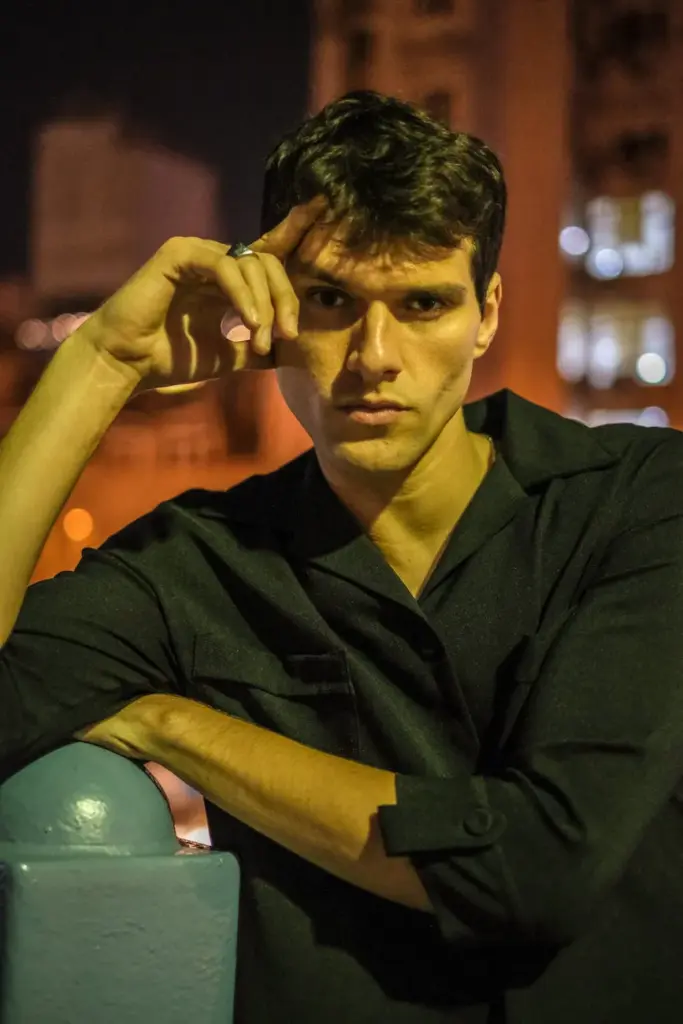 Stylish man in black shirt posing at night with city backdrop, exuding urban fashion vibe.