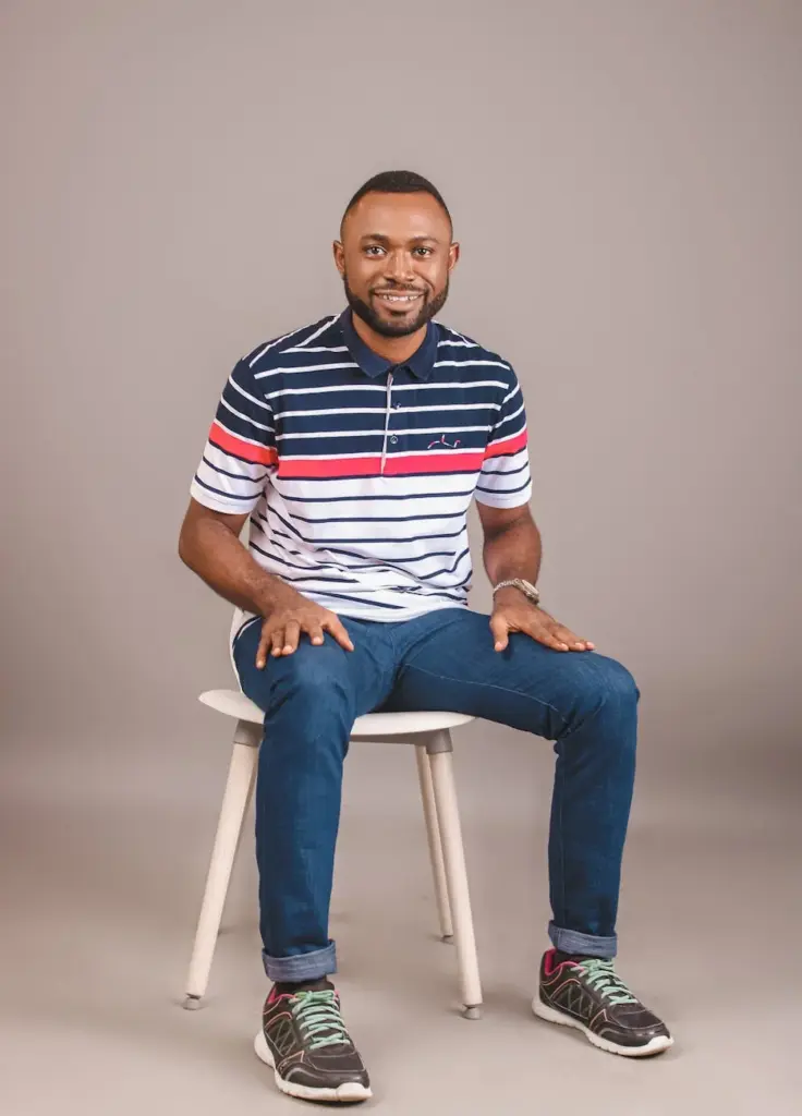 A cheerful man sitting on a stool in a studio wearing a striped polo shirt and jeans.