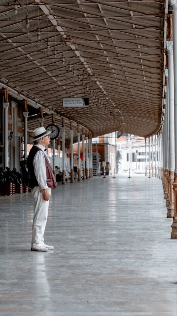 Man dressed in vintage attire standing at an empty railway station, reflecting travel and waiting themes.