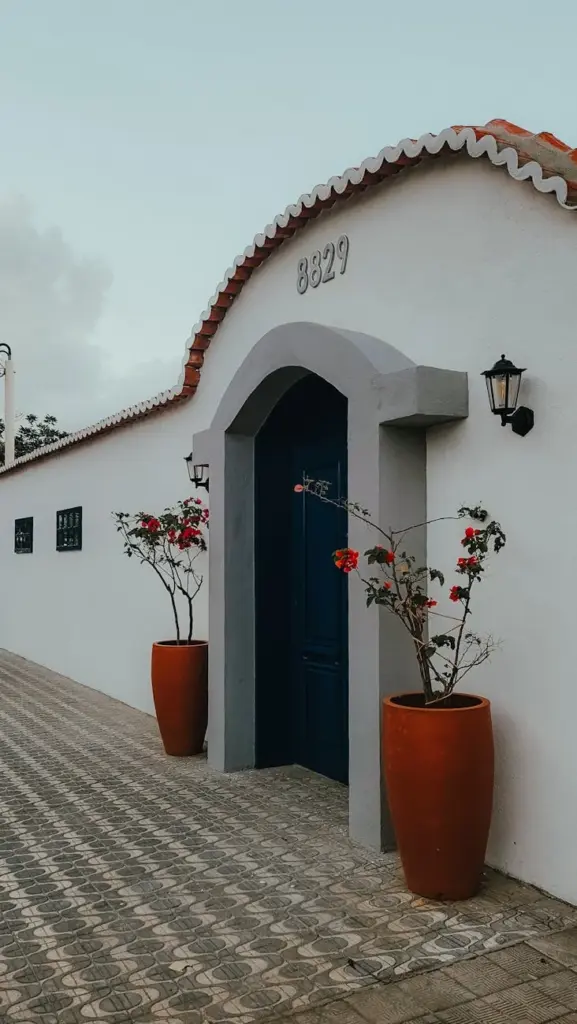 A quaint white house with a blue door and red tiled roof, featuring potted flowering plants and lanterns.