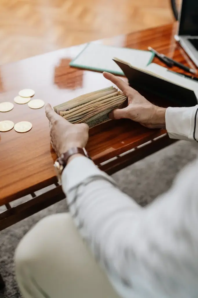 A person counting banknotes on a wooden desk indoors with coins nearby.