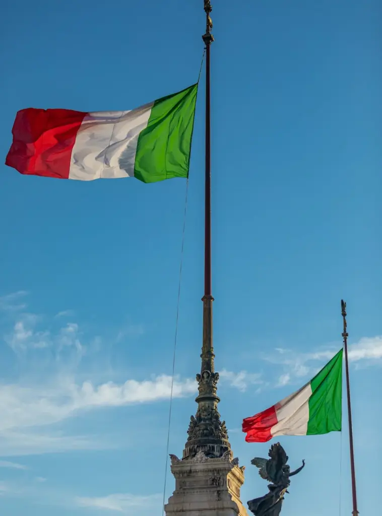 Italian flags waving at the Vittoriano monument in Rome under a clear blue sky.