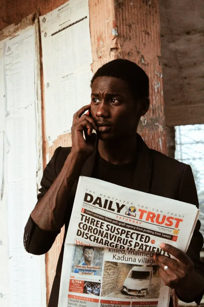 Young African American male in brown jacket standing with newspaper and talking by mobile phone in daylight