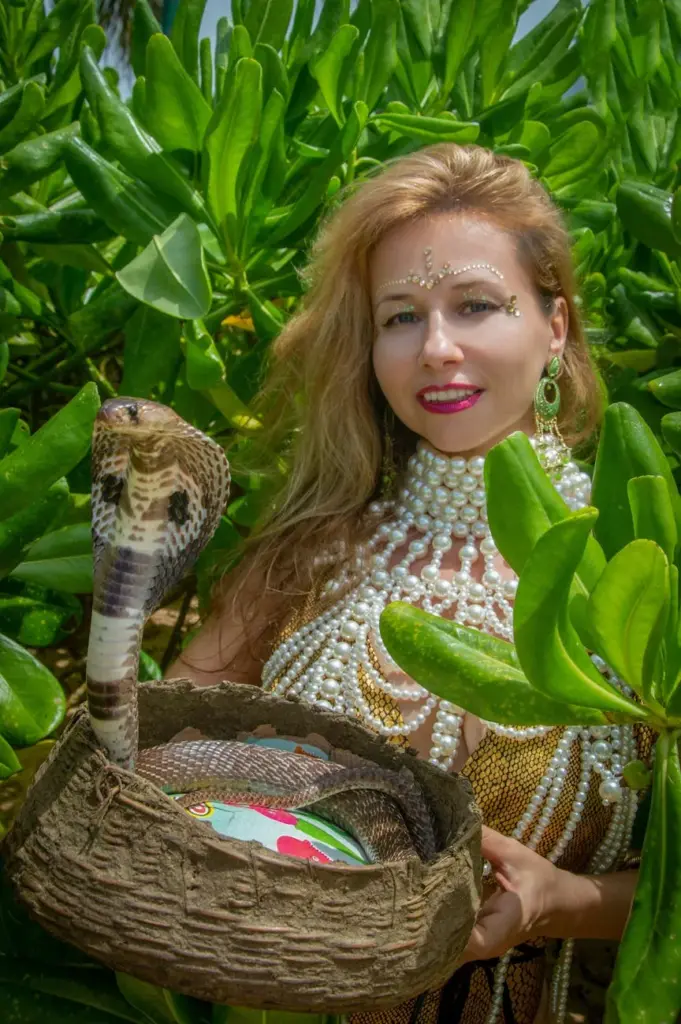 A woman holding a basket with a cobra, surrounded by lush greenery.