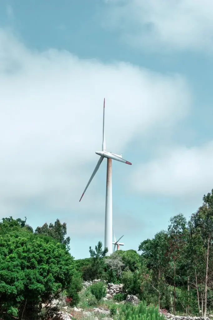 A towering wind turbine stands amidst lush greenery under a cloudy blue sky.