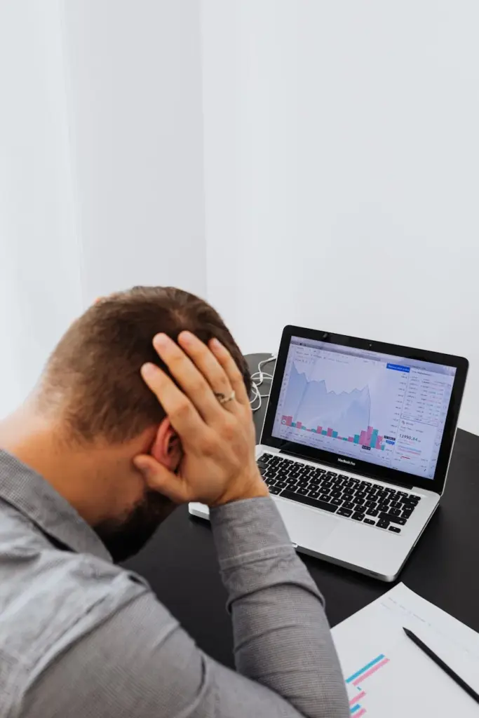 Businessman holding head in frustration, analyzing financial charts on laptop.