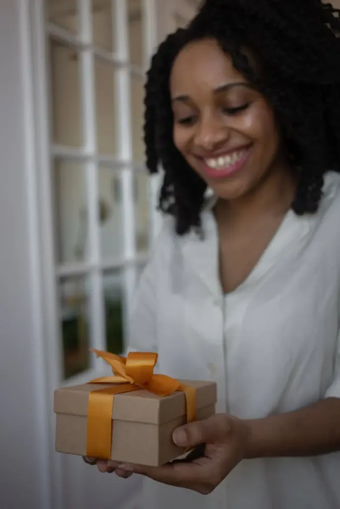 An African American woman joyfully holds a gift box wrapped with an orange ribbon indoors.