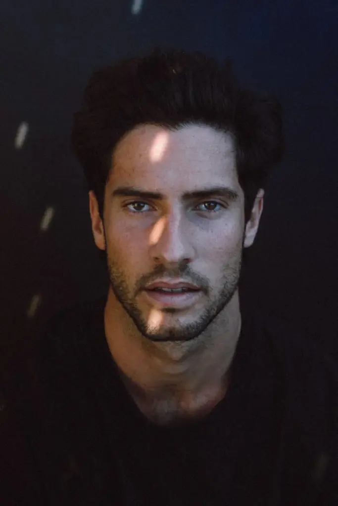 Handsome young pensive Hispanic male model with dark hair in casual clothes looking at camera with concentration in black studio with sun flares