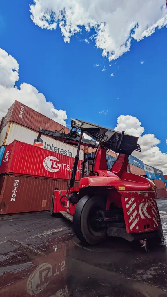 Dynamic view of a red forklift amidst stacked shipping containers under a vibrant blue sky.