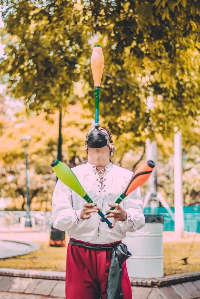 A talented street performer skillfully juggles colorful clubs in a sunny park setting.