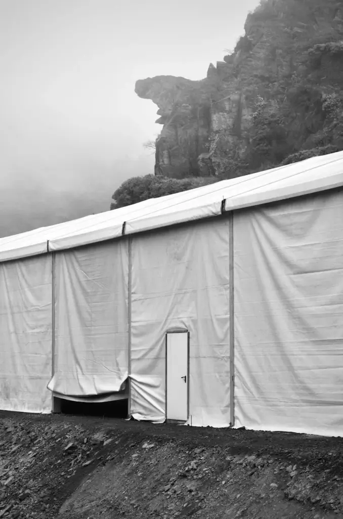 A large white tent stands against a rocky cliff, enveloped in fog, creating a dramatic monochrome scene.