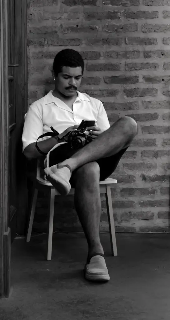 Casual portrait of a young man using a smartphone and holding a camera, seated against a brick wall in Buenos Aires.