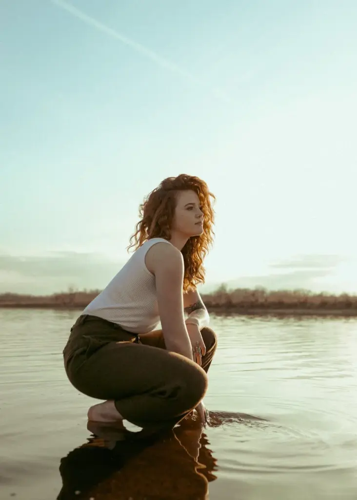 A young woman with curly hair crouches by a serene lake during sunrise. Captures peaceful summer vibes.