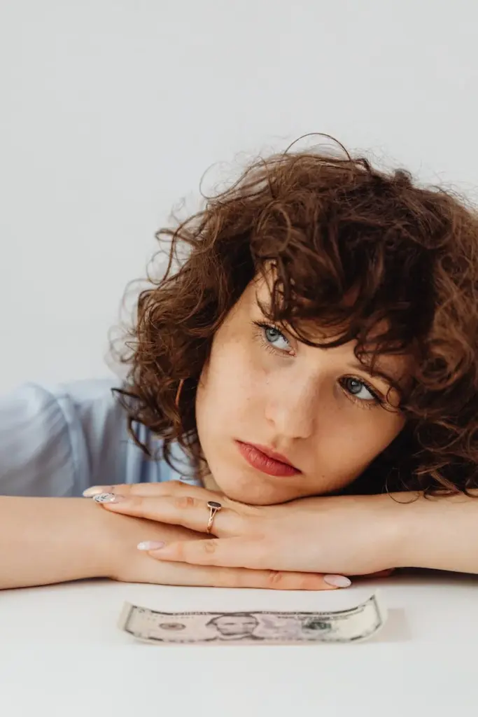 Woman with curly hair resting head on table, contemplating with dollar bill in view.