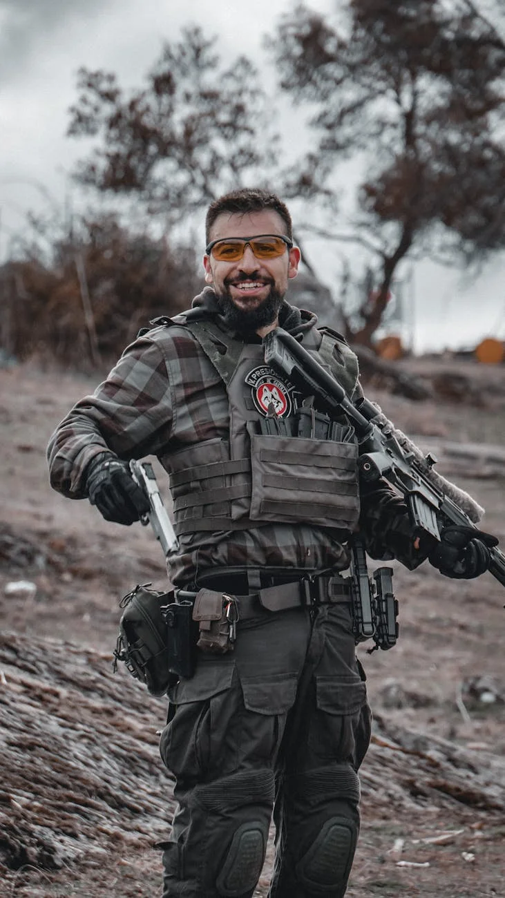 Smiling man in military uniform and tactical gear holding firearms outdoors.