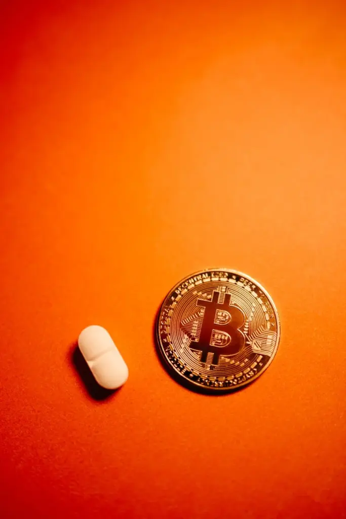 Bitcoin coin next to a pill on an orange background representing finance and health concepts.