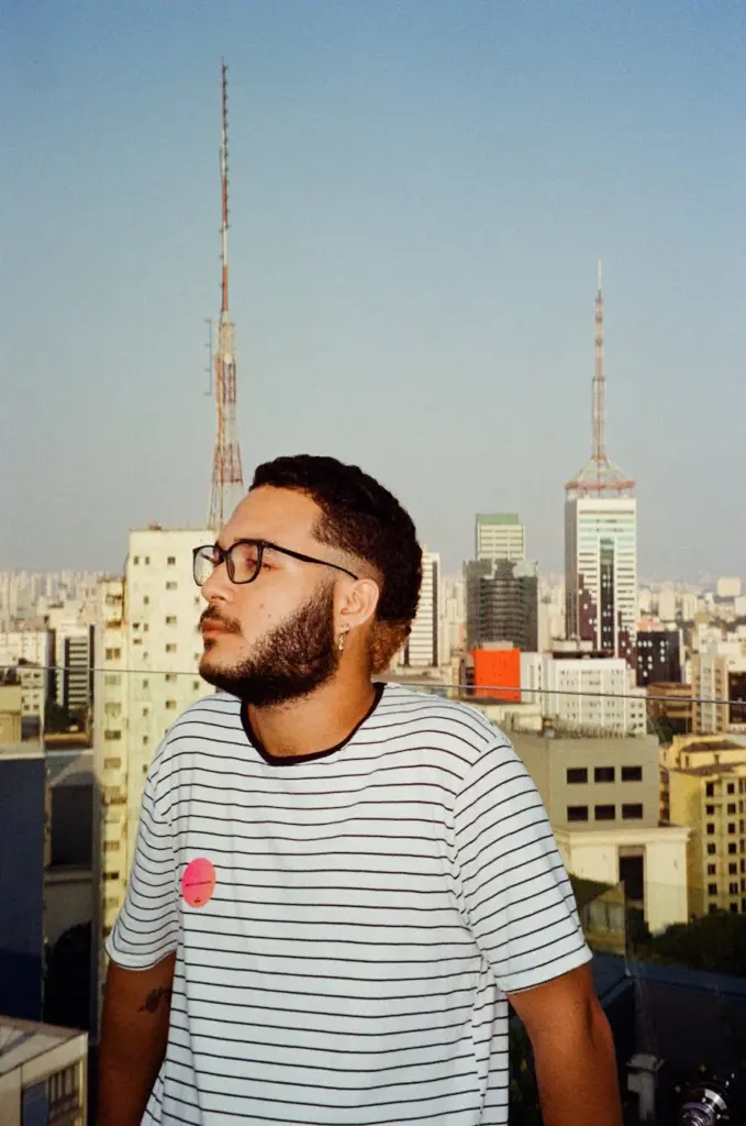 A young man with glasses poses against São Paulo's distinctive skyline on a sunny day.