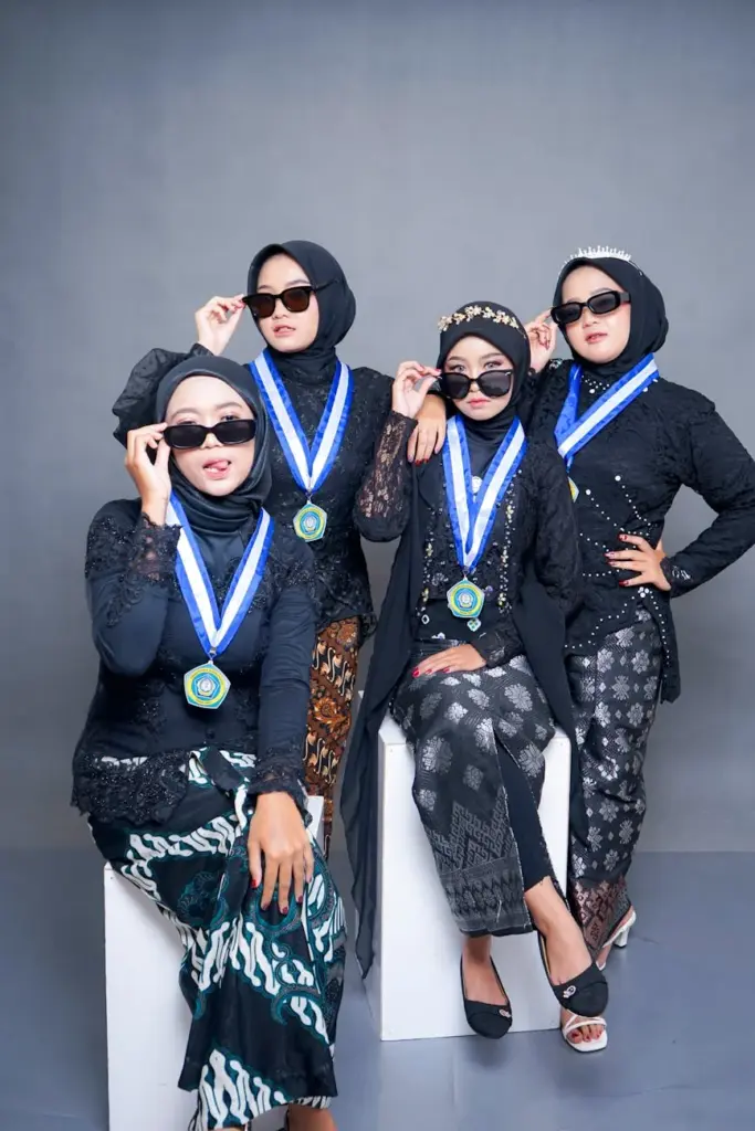 Four women in hijabs celebrate graduation with sunglasses and medals.