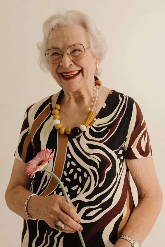 Joyful senior woman in patterned dress holding a pink gerbera flower and smiling warmly.