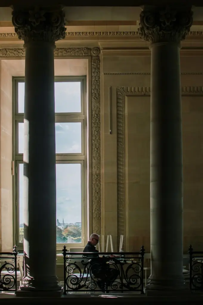 A man sits reading between grand columns in a historic Parisian building with a view of the city through large windows.