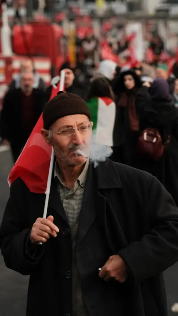 Elderly man smoking and holding a flag during a protest. A crowd in the background.