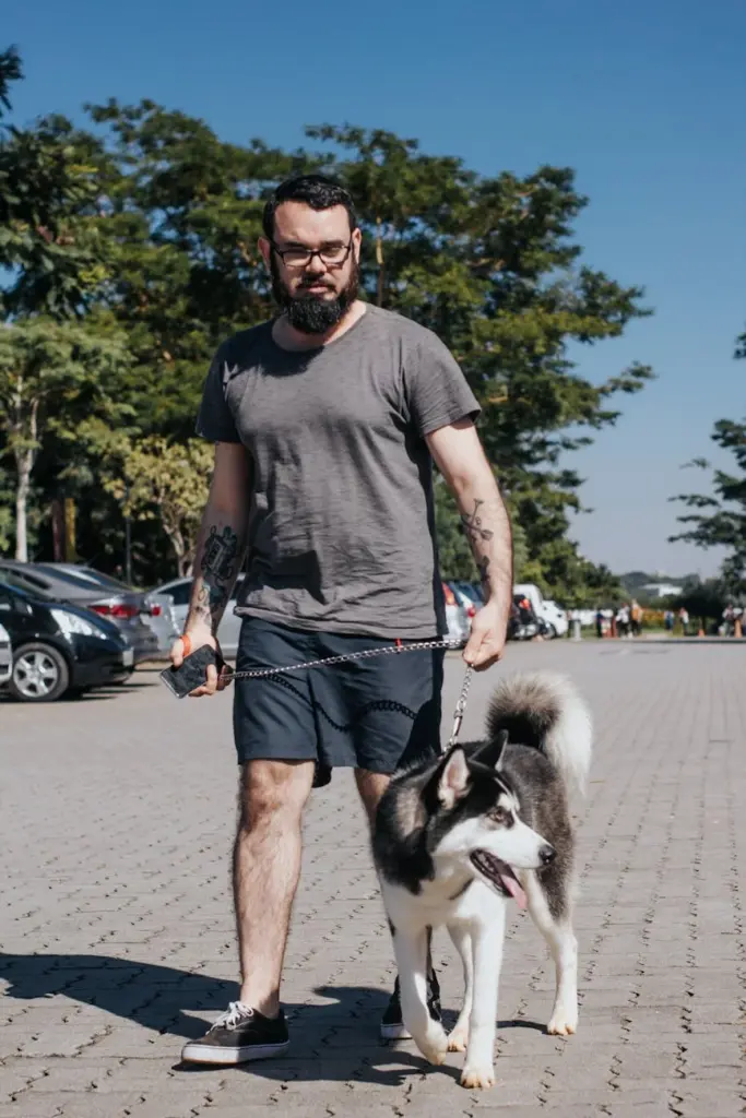 A bearded man walks a Siberian Husky outdoors in a sunny park setting.