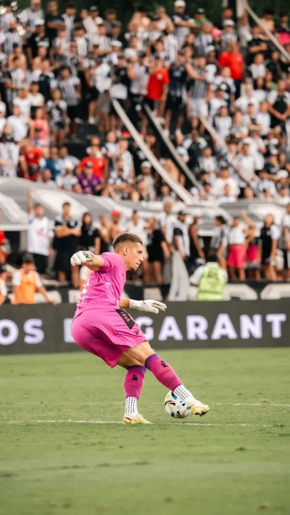Goalkeeper in pink kit kicking ball during a soccer match in crowded stadium.
