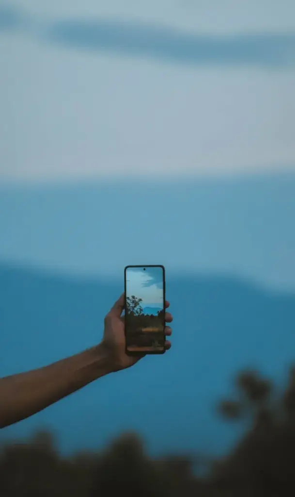 A hand holding a smartphone capturing a mountain landscape during the day.