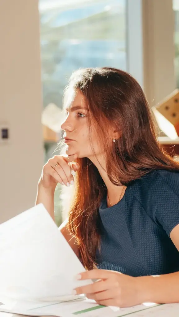 A thoughtful woman looks out the window while holding papers, bathed in warm sunlight indoors.
