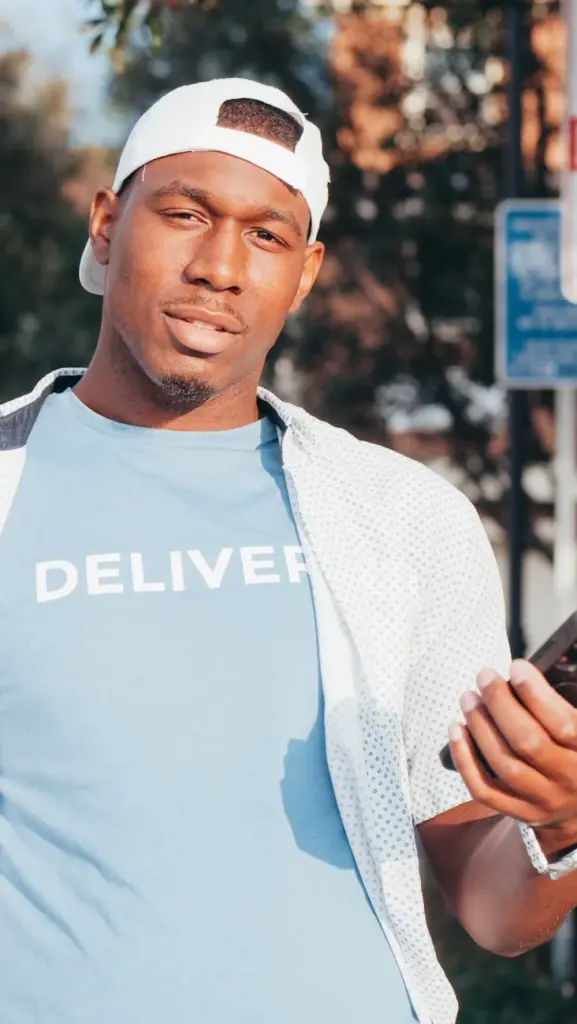 Casual delivery person holding a smartphone outdoors, wearing a 'Deliver' shirt and cap.
