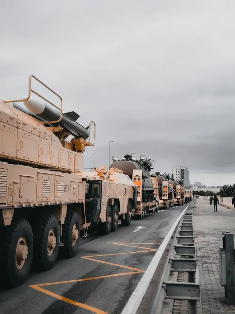 A line of military vehicles travels along an urban road under a gray, overcast sky.