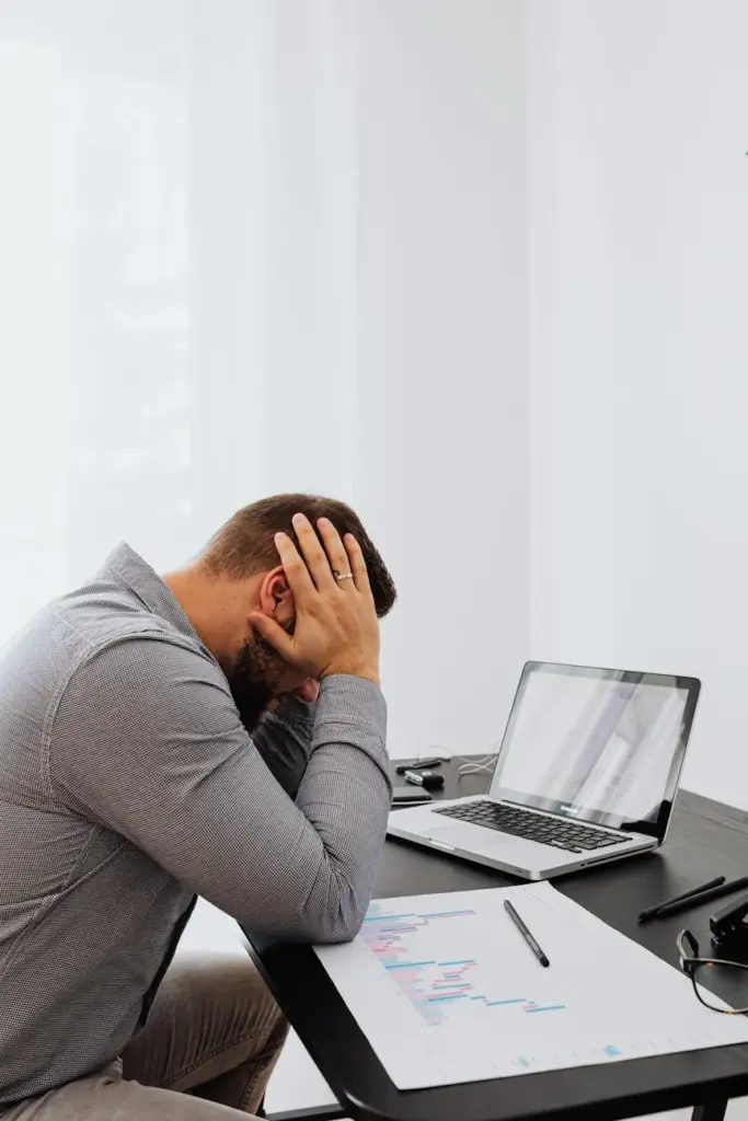 A frustrated man sits at a desk with a laptop and charts, showing work stress.