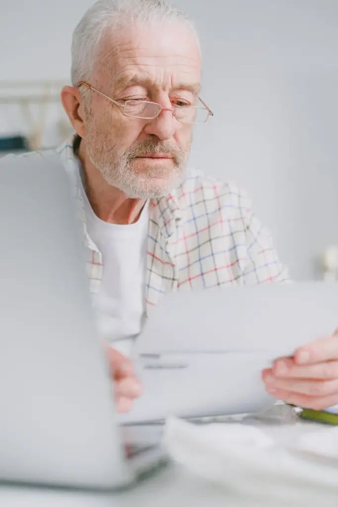 Elderly man with glasses reviews paperwork while sitting indoors with a laptop.