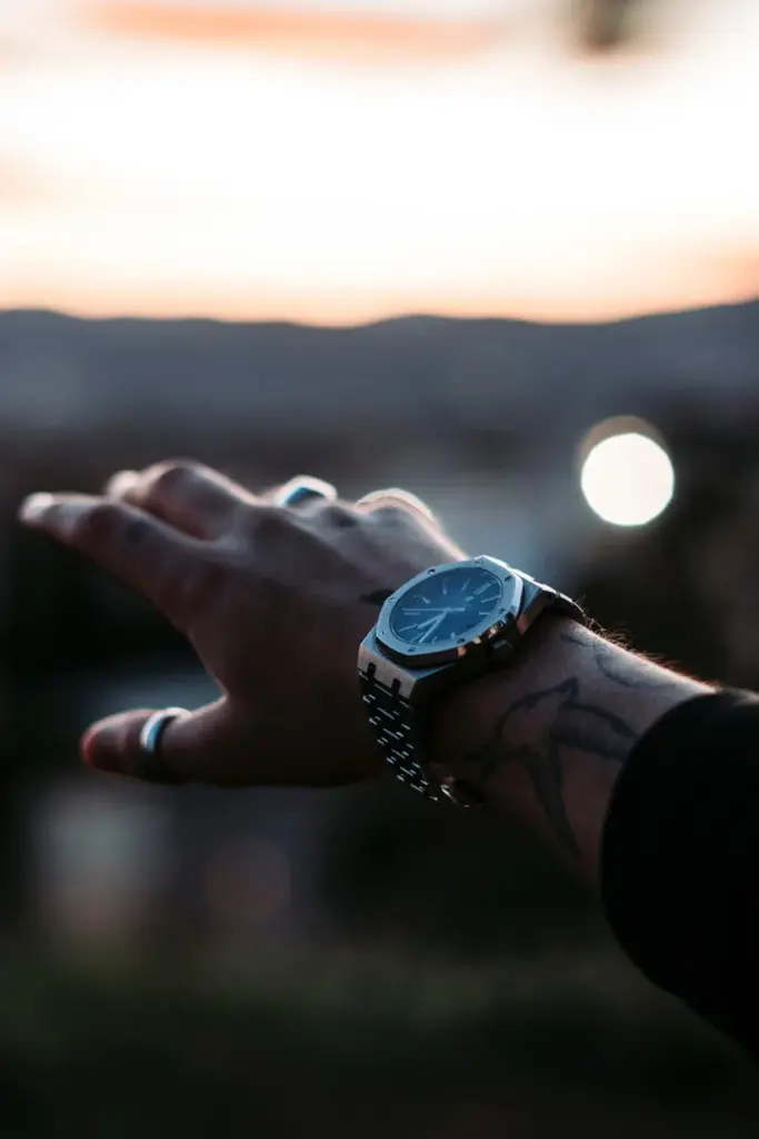 A hand with tattoos reaching out, adorned with a wristwatch against the backdrop of a sunset.