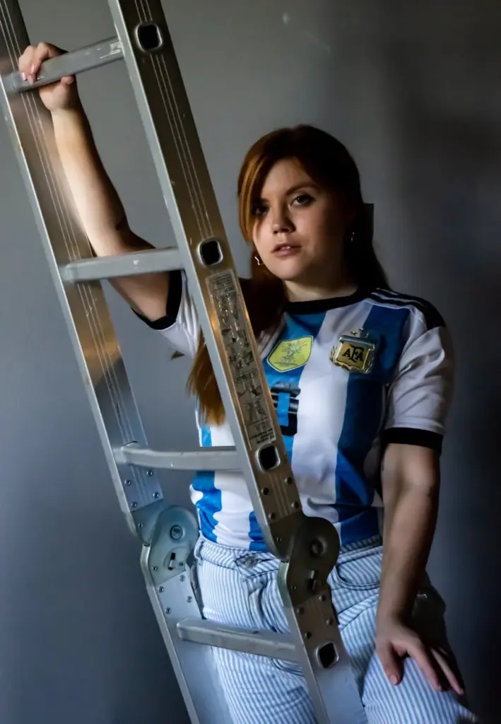 Adult woman in Argentina soccer jersey poses with a ladder indoors.