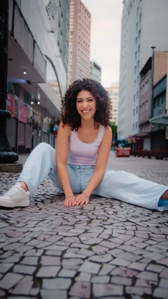 Confident young woman smiling while sitting on a city street, showcasing urban fashion.