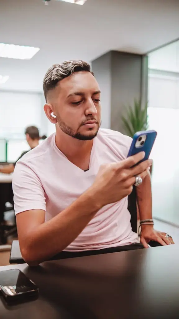 A young man with short hair uses his smartphone in a modern office setting, sitting at a desk.