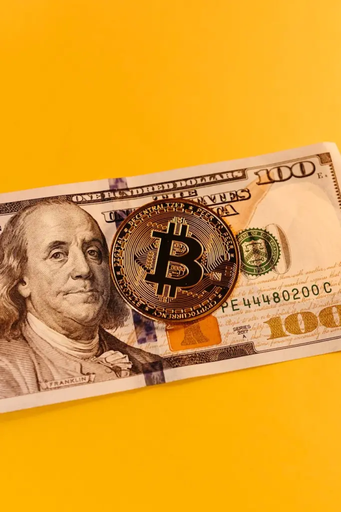 A Bitcoin coin placed on a US one hundred dollar bill against a yellow background, symbolizing cryptocurrency and traditional currency.