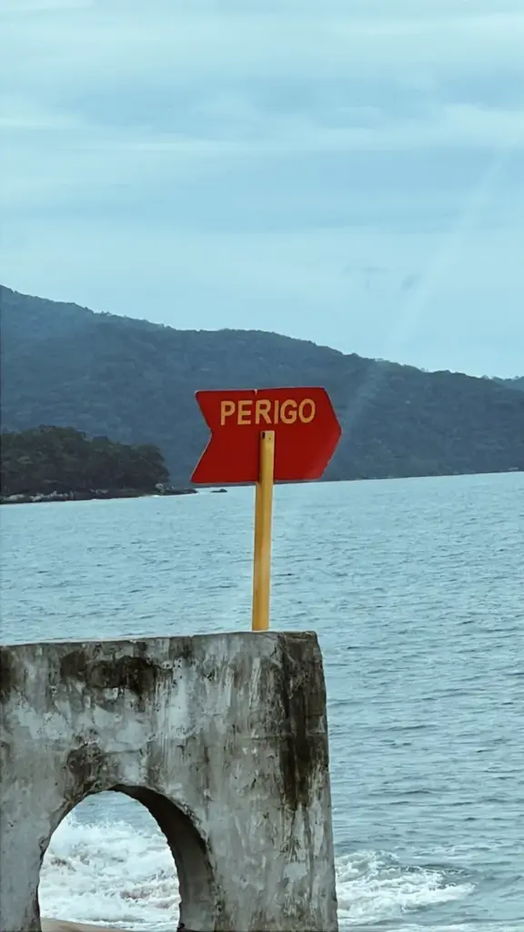 Red 'Perigo' warning sign on a pier in Caraguatatuba, Brazil, with ocean backdrop.