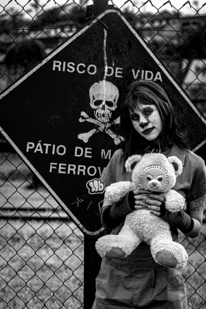 Artistic black and white portrait of a woman in costume holding a teddy bear by a danger sign.