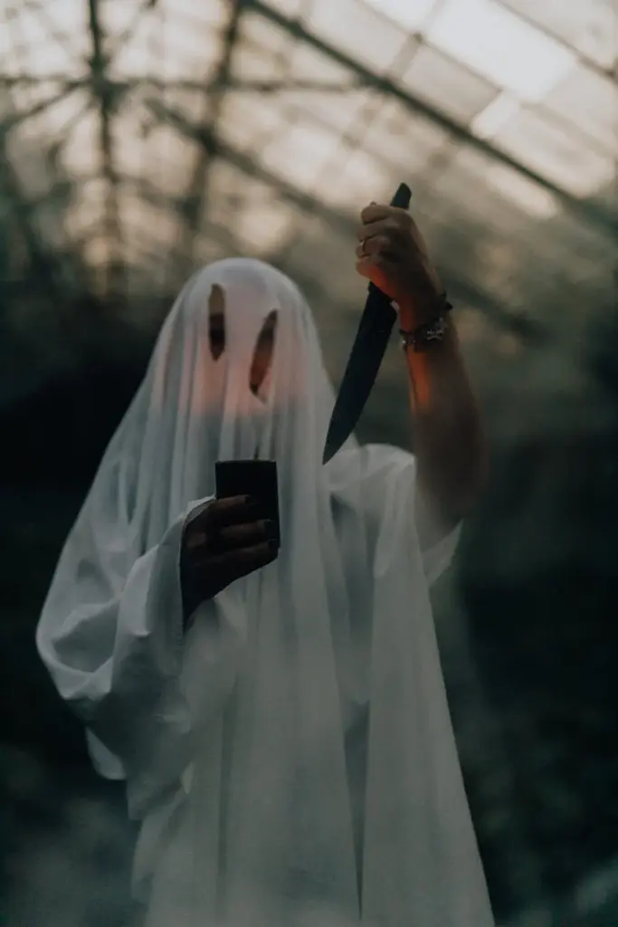 Creepy ghost costume holding a knife inside a dimly lit greenhouse, perfect for Halloween themes.