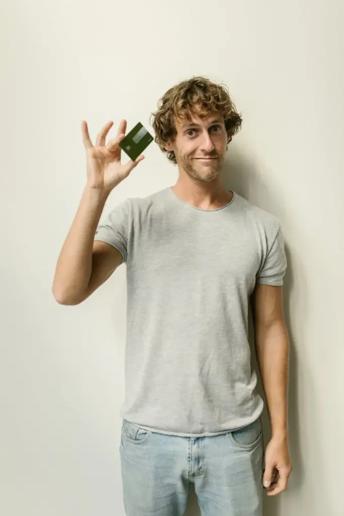 Caucasian man smiling while showing a green card, standing against a white background, wearing a gray shirt.