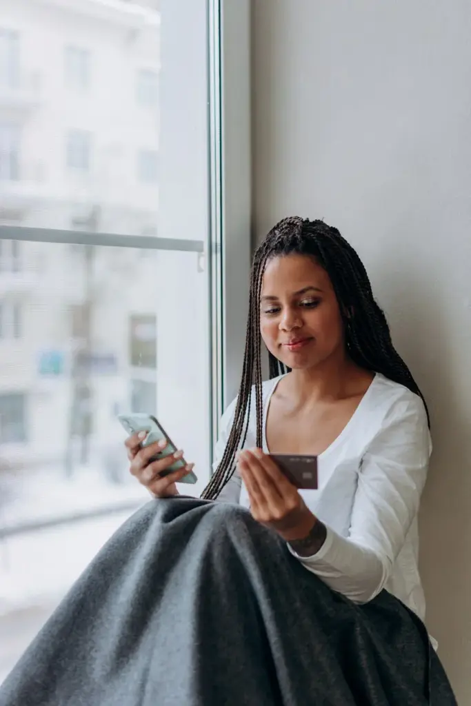 A woman using a smartphone and credit card for online shopping by a window.