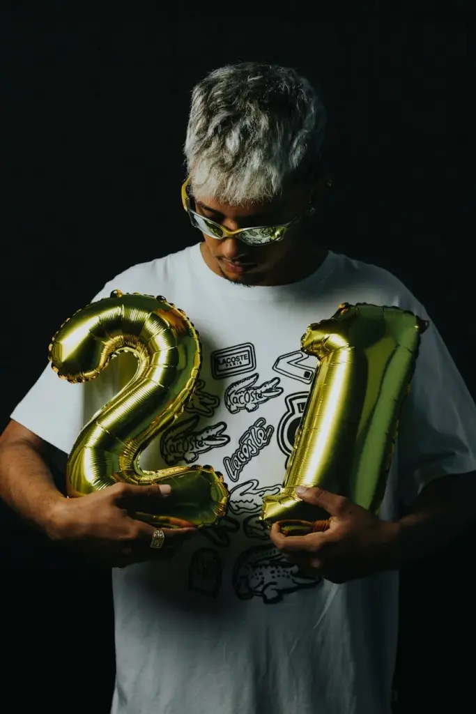 Portrait of a stylish man holding gold '21' balloons against a black background, celebrating his 21st birthday.