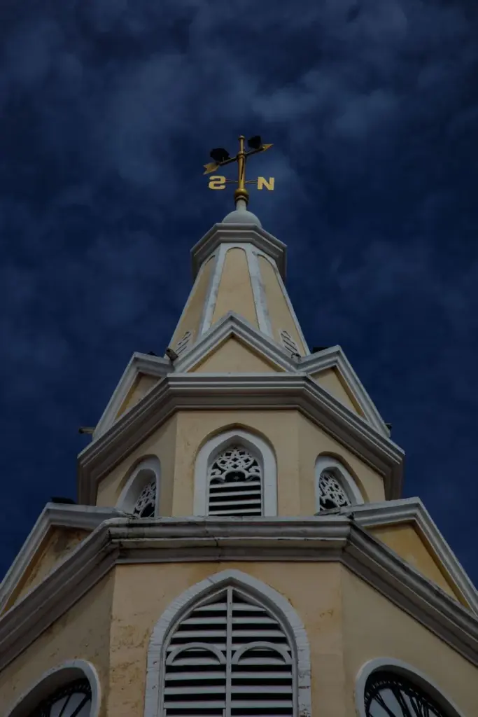 Upward view of a historic church steeple with a weather vane against a dramatic blue sky.