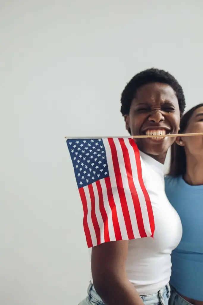 A smiling woman holding a small American flag indoors, celebrating joyfully.