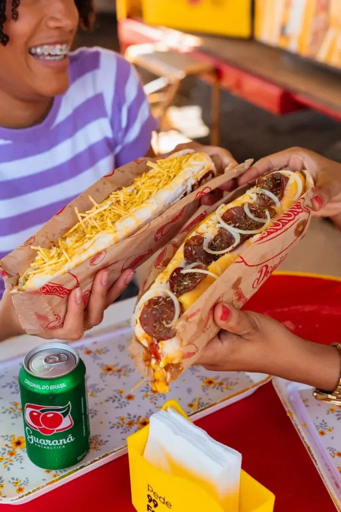 Two friends sharing Brazilian-style hot dogs and a Guaraná drink at an outdoor setting.