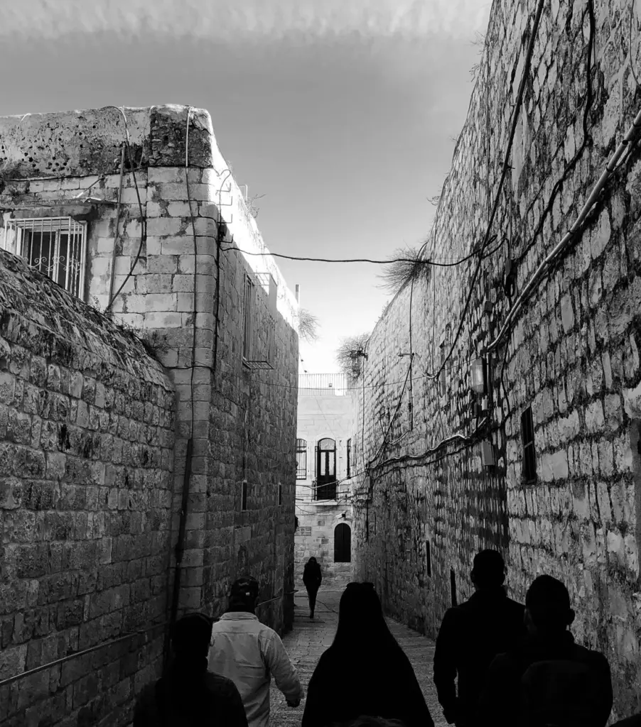 A monochrome photograph of people walking down a narrow alley in historic Jerusalem.